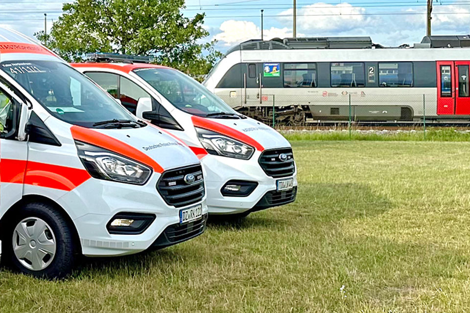 Zwei Krankentransportwagen stehen nebeneinander auf einer Wiese. Im Hintergrund steht der S-Bahn-Zug auf dem Hauptfahrgleis Bitterfeld Richtung Leipzig.