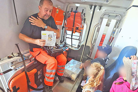 Im hinteren Teil eines Krankentransportwagens sitzt ein Rettungssanitäter auf der Krankentrage. Ihm gegenüber stehen drei kleine Kinder und schauen auf die Beschreibungstafel in seinen Händen.